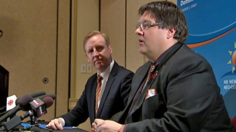 Two men sitting beside each other with a cluster of microphones in front of them.