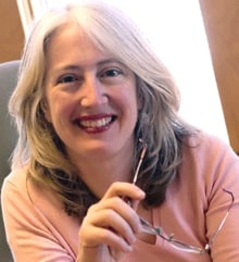 A woman with grey hair wearing a pink shirt holding her glasses