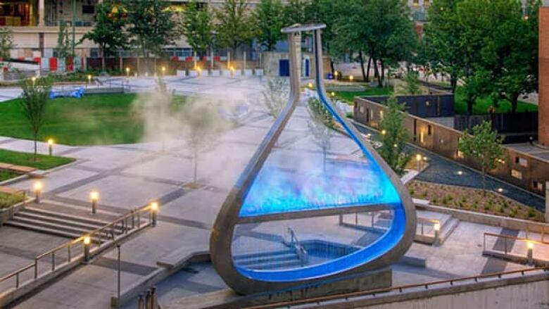 A flask-shaped piece of illiminated public art within a concrete plaza.