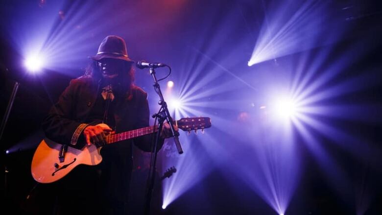 A man with a guitar stands in front of bright stage lights. 