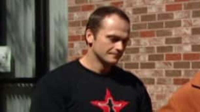 A man in a black T shirt is shown against the backdrop of a brick building.