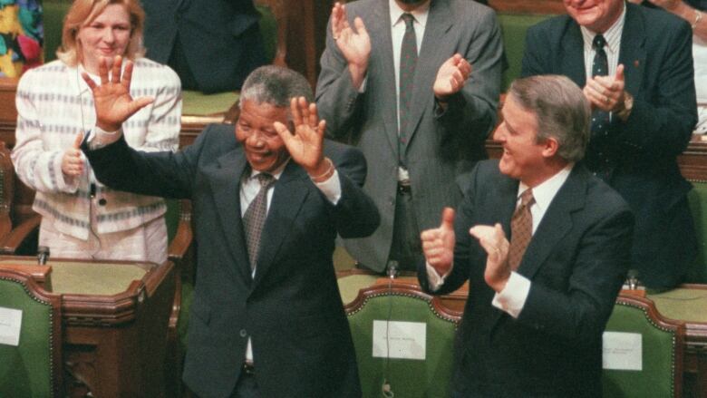 A man in a suit stands and waves with both hands in the air as others around applaud him.