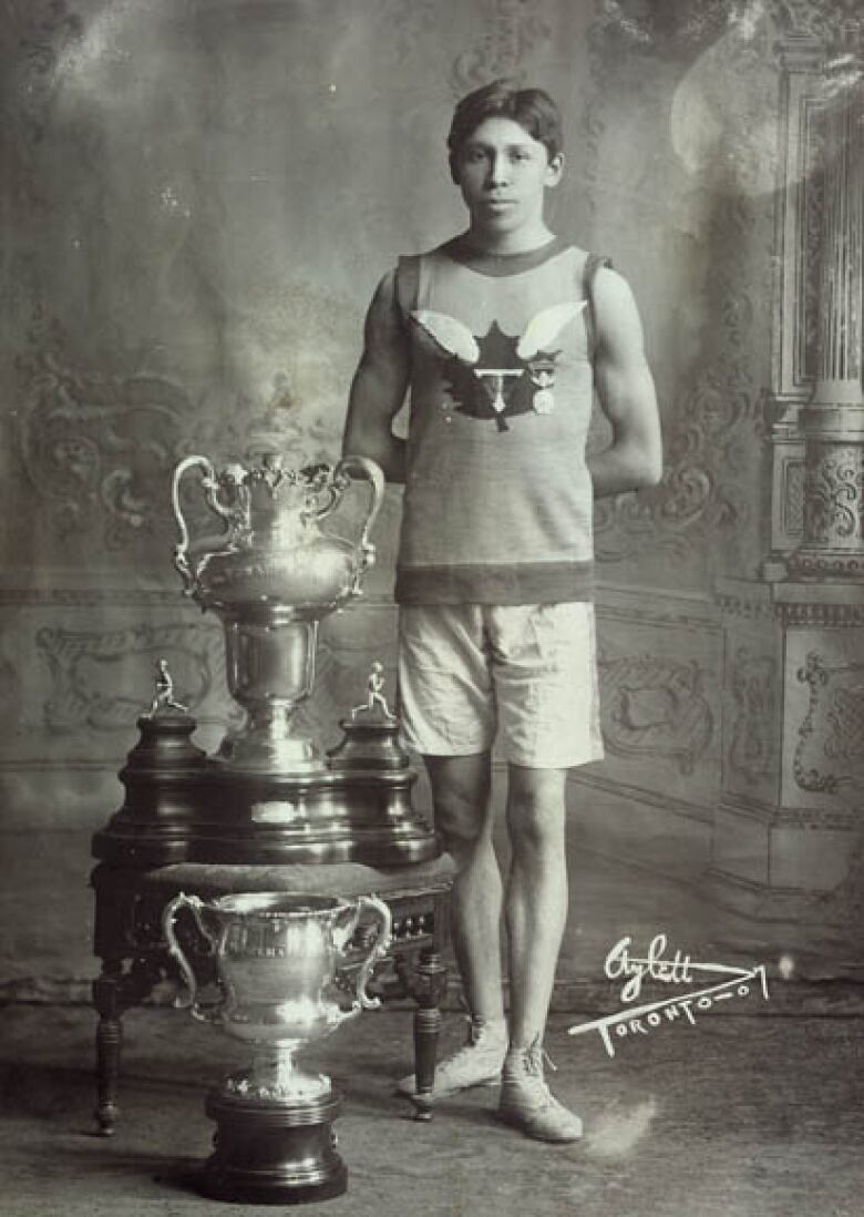 In a black-and-white photo, Tom Longboat stands next to a trophy.