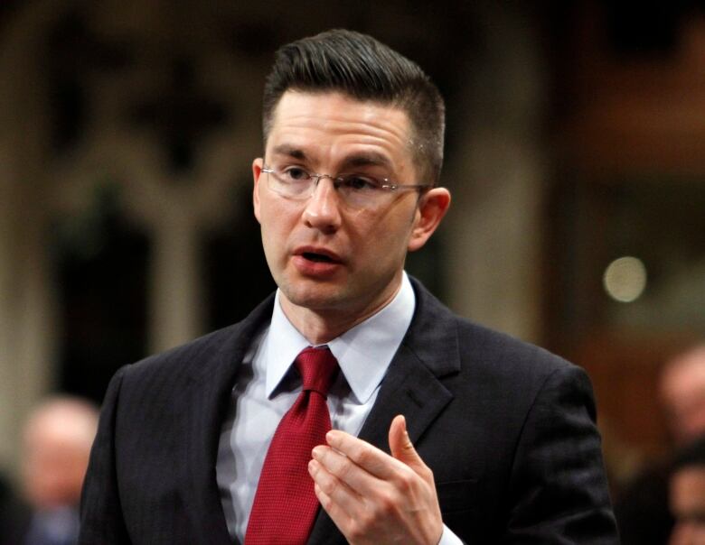 A man with glasses, wearing a dark suit and a red tie, stands to speak.