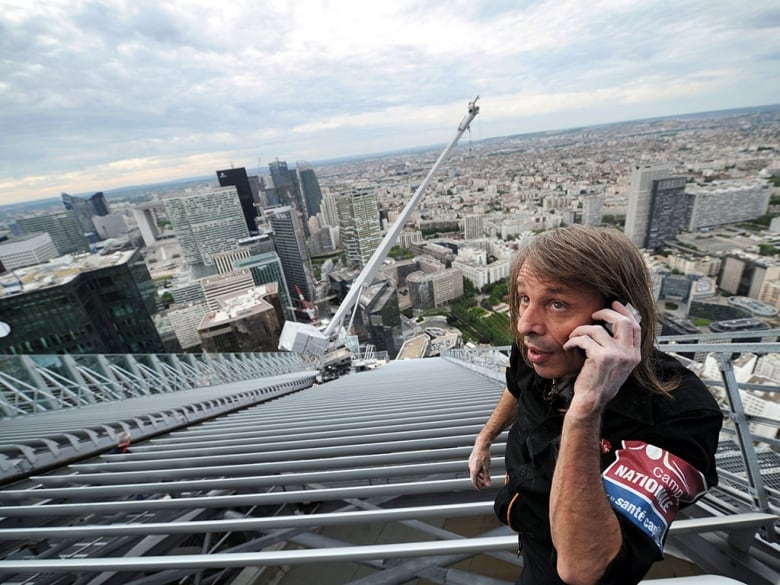 A man pictured from above talking on a cellphone while perching high up on the side of a skyscraper. 