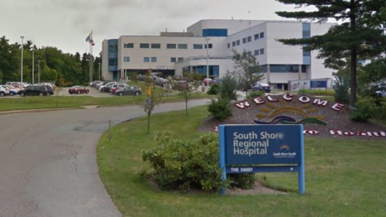 A blue sign reads South Shore Regional Hospital by the driveway leading to a large white and blue hospital, with cars in a parking lot