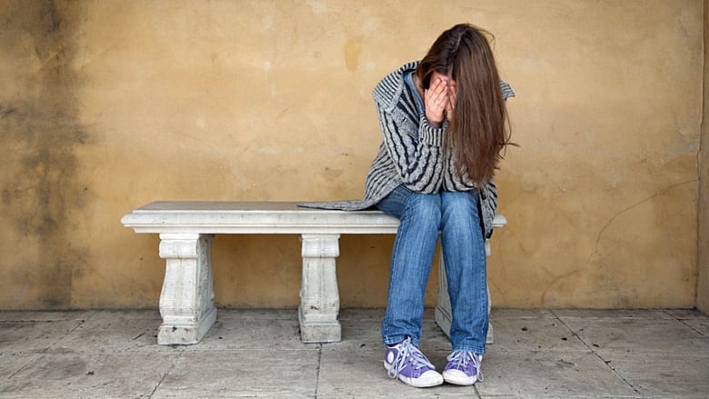 A young woman with long hair, wearing jeans and sneakers, sits on a bench with her face in her hands, as if in despair. 