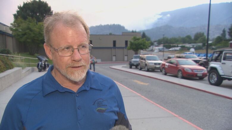 A white-haired man in a blue T-shirt with glasses stands in front of a traffic road.