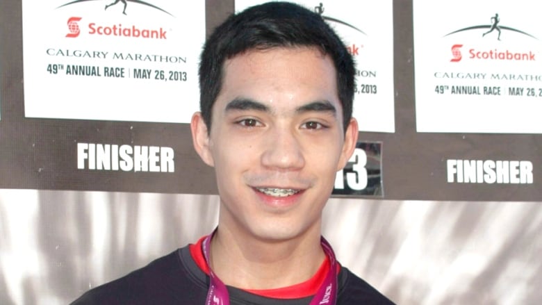 A young man with dark hair smiles while holding up a medal around his neck that says 