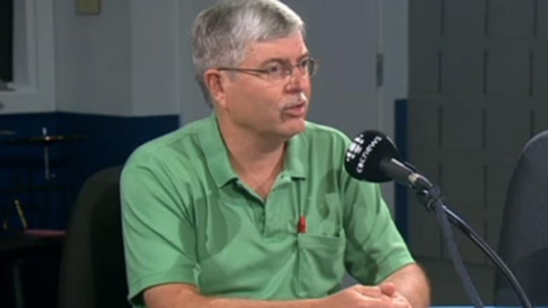 A silver-haired man in glasses and a green polo shirt sits and speaks into a CBC News microphone.