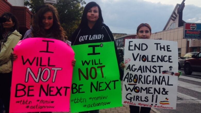 Three girls standing on a street holding signs. 