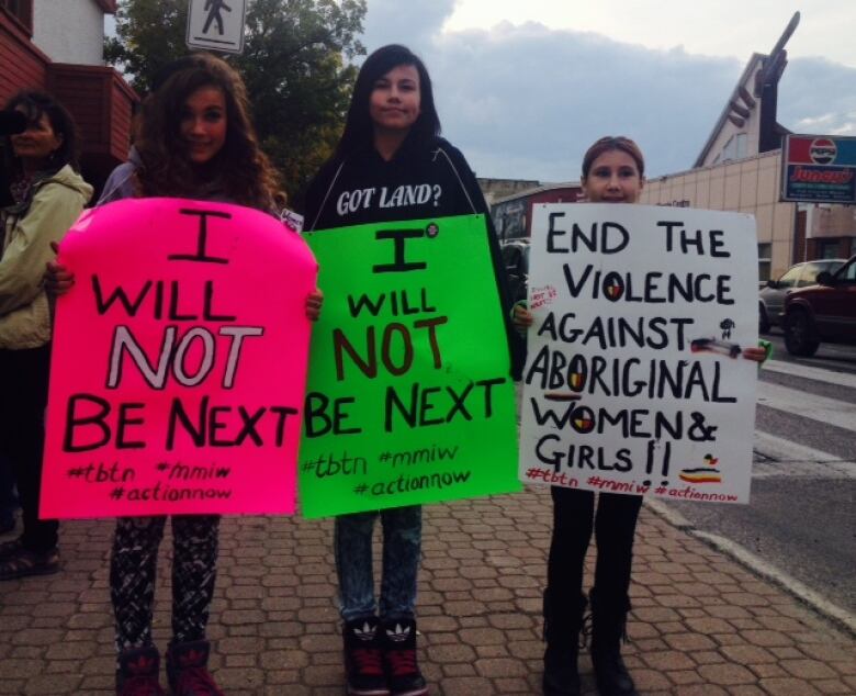 Three girls standing on a street holding signs. 