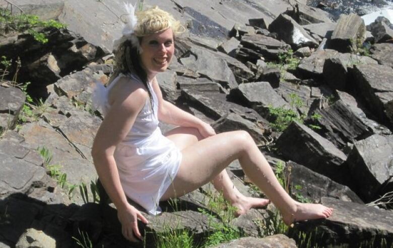 A woman wearing a white dress sits on a rock pile near the ocean. 