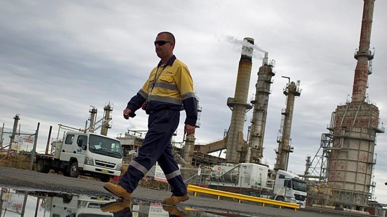 A worker in a yellow jacket and work boots walks in front of an oil refinery.