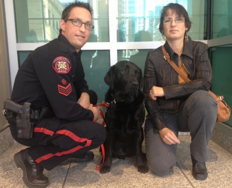 A black lab sits between a police officer and a woman.