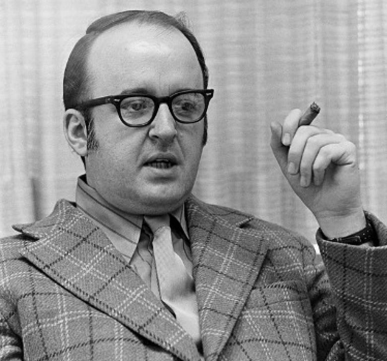 A bespectacled cleanshaven and balding man is shown holding a cigar in his left hand while wearing a striped suit and a tie in a black and white photograph that appears several years old.