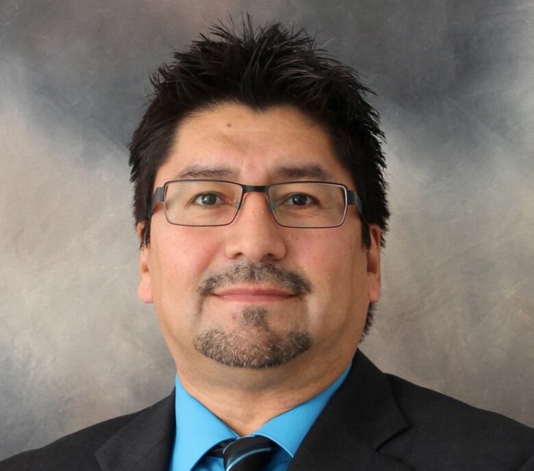 A headshot of a man with dark hair and a blue dress shirt and dark tie.