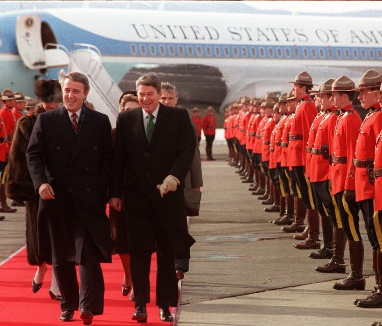 Two men walk down a red carpet from an airplane as a line of men in red uniforms stand at attention.