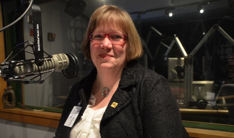 A smiling woman with a blond bobbed haircut, pendant necklace, white blouse and dark gray jacket sits in a radio studio with microphones.