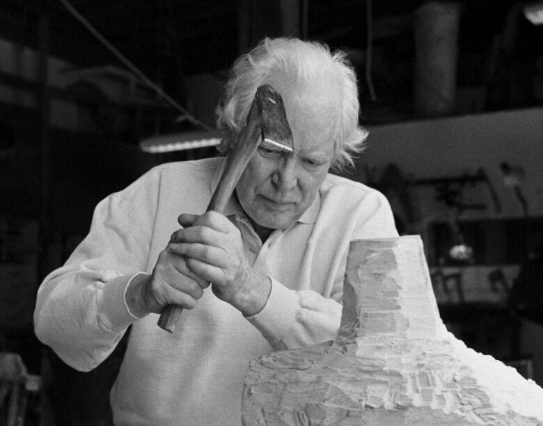 A black and white photo of a man with grey hair carrying a hammer as he carves a sculpture. 