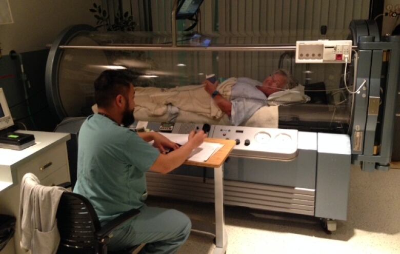 A woman lies in an enclosed bed in a hospital setting with a health care working sitting next to the machine.