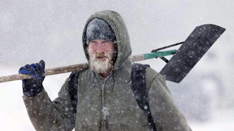 A man with a frosted beard stands in a snow storm holding a shovel