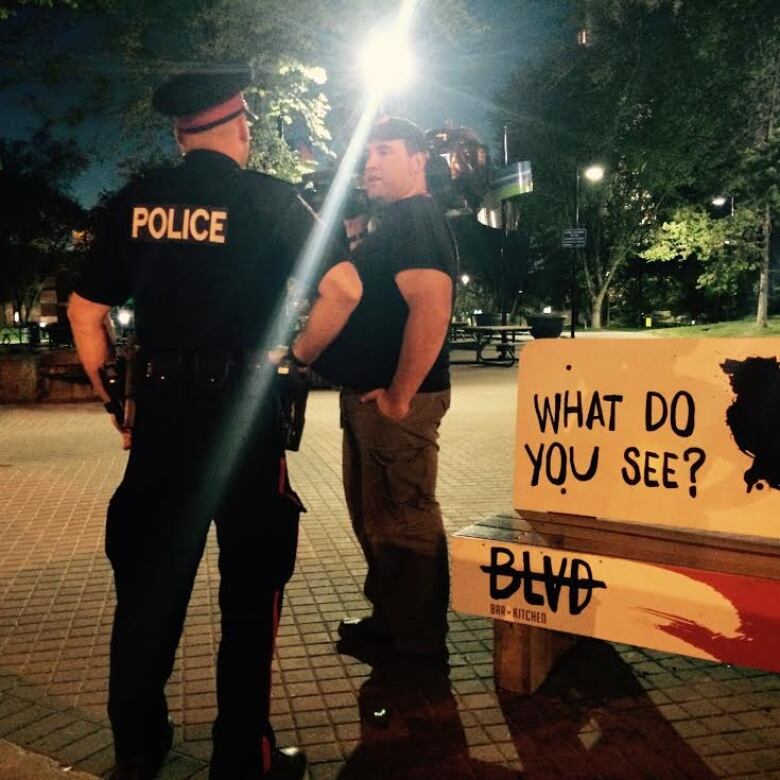 A police officer talks to civilian near a bench at night time. 