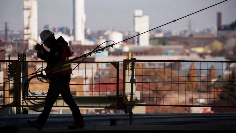 A person is walking in silhouette with a hard hat on in the left of the frame. Tall buildings can be seen in the background. 