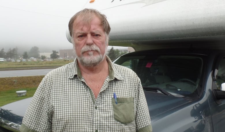 An older man in a checked shirt stands in front of a truck.