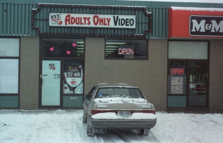 A grey 1990s sedan is parked in a snowy parking lot of a strip mall in front of a store with a sign that reads 'Adults Only Video.' 