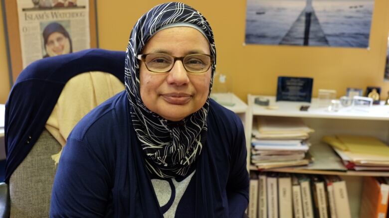 A seated woman wearing hijab leans forward and looks at the camera.