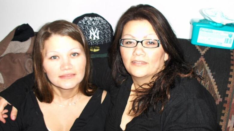 Two women, both with dark hair and dark shirts, sit on a couch together and look into the camera.