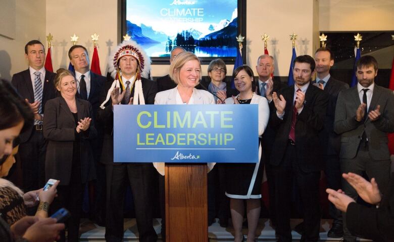 Rachel Notley, smiling, stands behind a podium that says 'Climate Leadership,' with a number of industry, environmental and Indigenous leaders clapping.