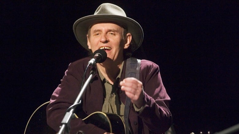 Man in a hat stands with a guitar and sings into a microphone. 