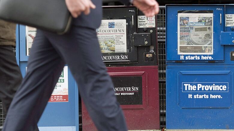 A closeup of a person's legs walking past three newspaper boxes on a city sidewalk.