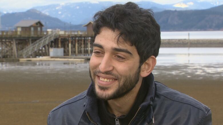 A Syrian man smiles at the camera in front of a body of water. He wears a black jacket.