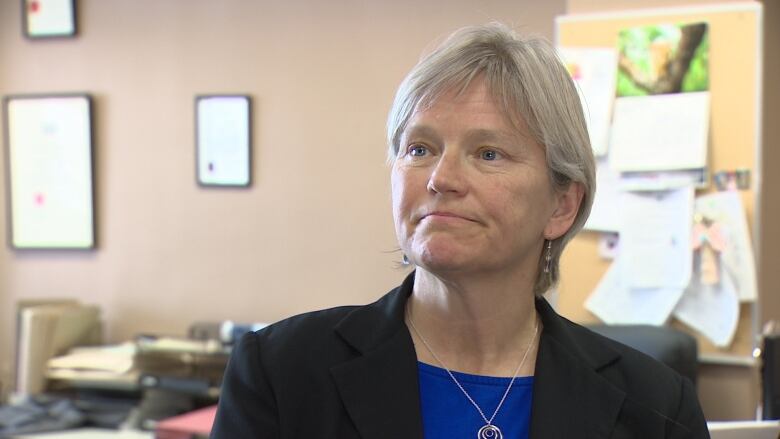 A woman wearing a suit jacket and a necklace is in an office setting.