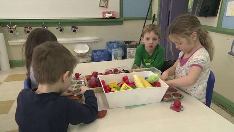 Four young children are seen sitting around a small table with a basket of toys in the middle.