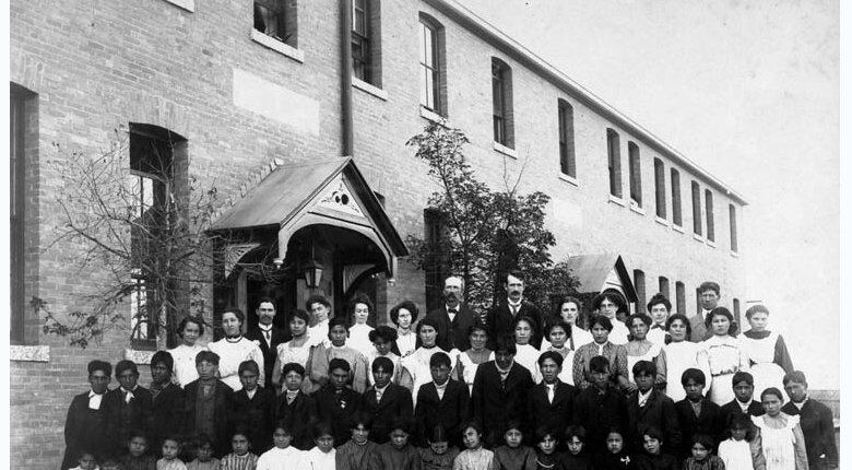 Children in old clothing stand in front of an old school.