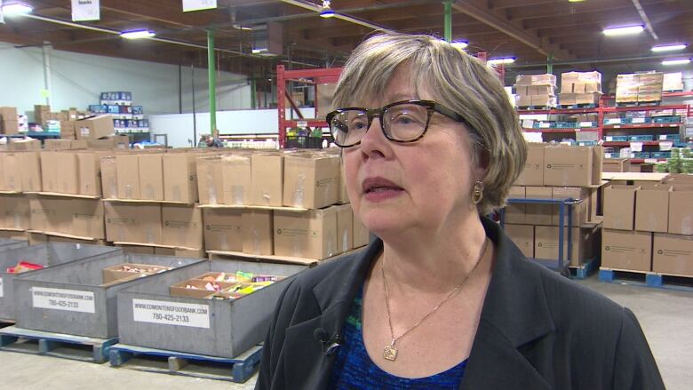 A woman wearing eyeglasses stands inside a warehouse, with bins of food in the background.