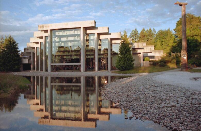 A modern-looking glass and concrete building is reflected in the water that fronts it.