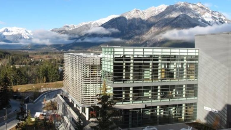 A large glass building is seen with trees, mountains and clouds in the background.