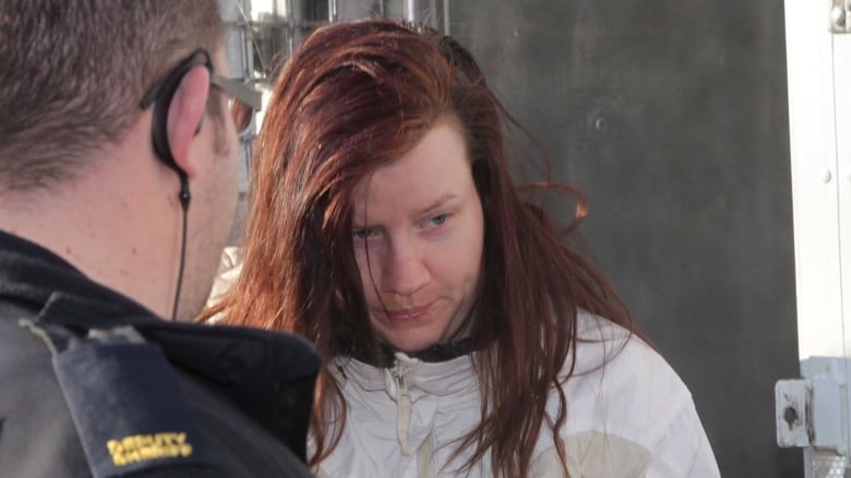 A white woman with dyed red hair is seen wearing an off-white jacket. A man wearing a police jacket stands in front of her.