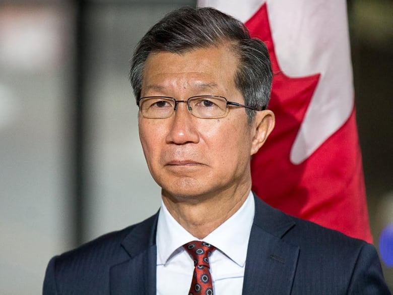 A man in a blue suit and glasses stands in front of a Canadian flag.