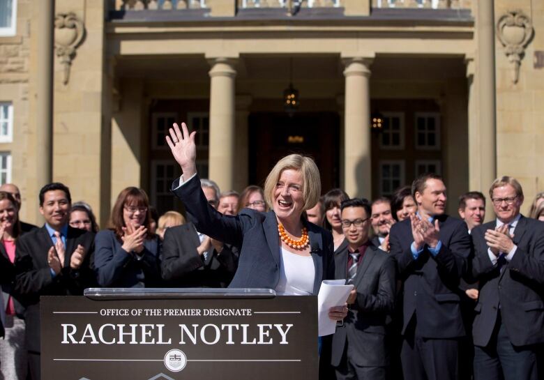 woman behind lectern smiles and waves, as fellow politicians gather behind her