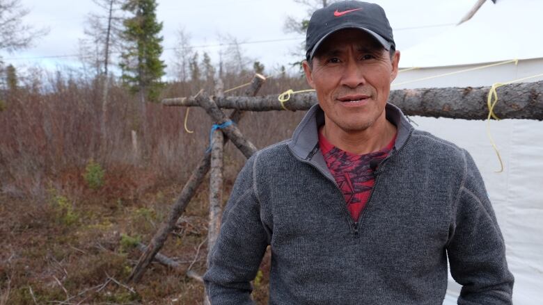 Man in sweater and hat stands in front of water and bush.