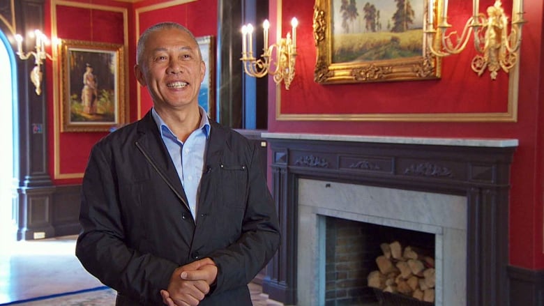 Mingfei Zhao smiles and clasps his hands in an opulent room with a fireplace and red walls.