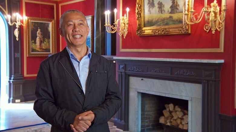 Mingfei Zhao smiles and clasps his hands in an opulent room with a fireplace and red walls.