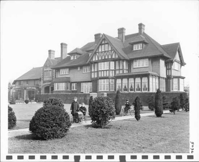 An archive black-and-white image of a large mansion, with a mother and child in the foreground.