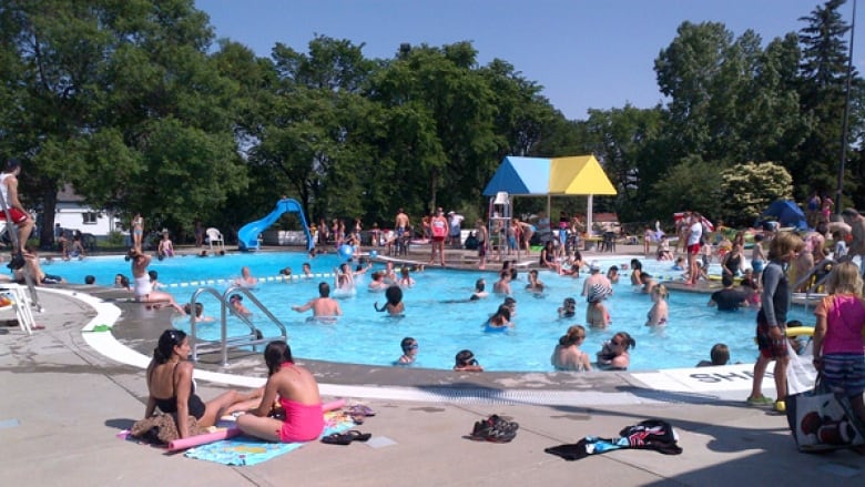 An outdoor pool is full of swimmers and young people wading in the shallow end on a sunny day.